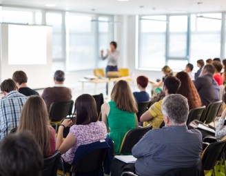 A group of people sat on chairs facing a lady who is talking and pointing at a board